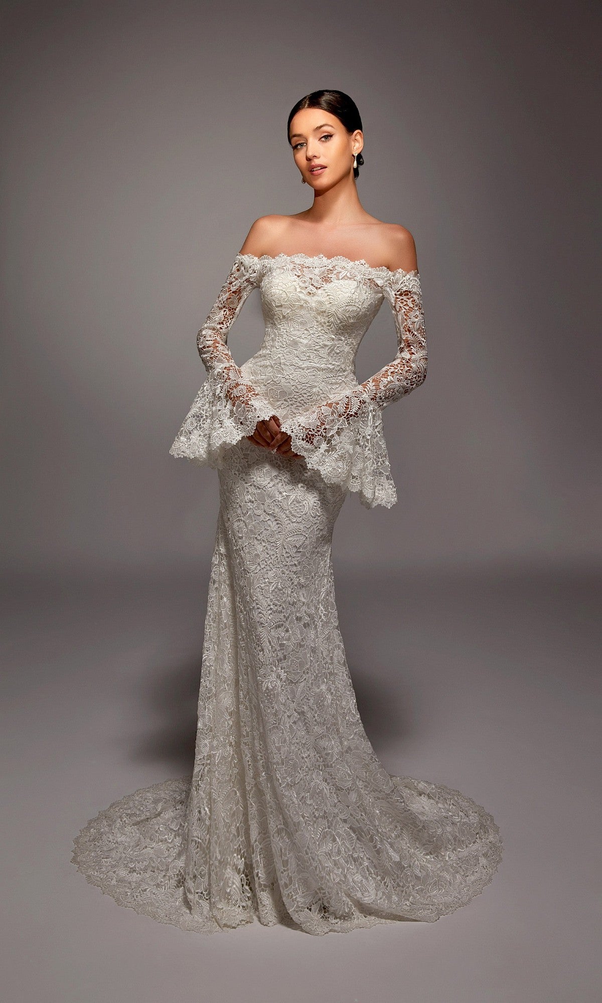 A woman stands in a studio with a neutral background, wearing the Long White Dress: Alyce 7156—an off-shoulder bridal gown featuring long lace sleeves.